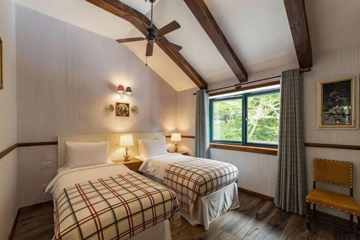 Una habitación con dos camas, ventilador de techo, ventana con vistas y un banco.