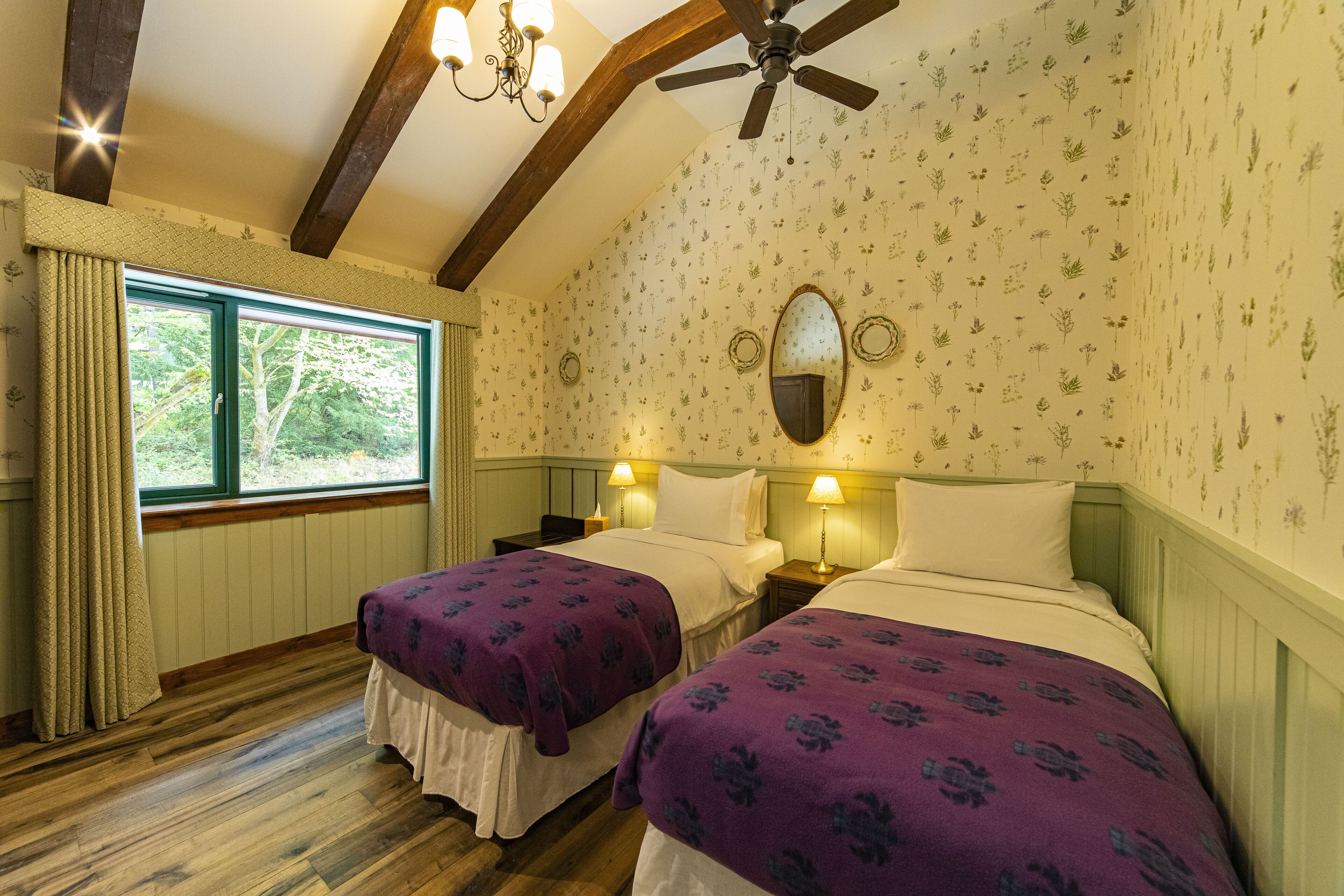 A room with two beds, wooden floors, a ceiling fan, and a window with curtains.