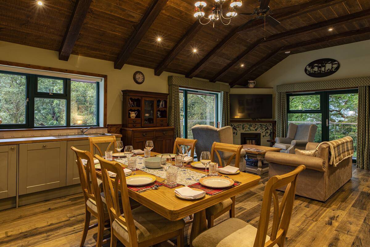 Un comedor con una mesa de madera dispuesta para cuatro personas, rodeada de sillas de madera. Se aprecia una zona de cocina con estufa y armarios al fondo.