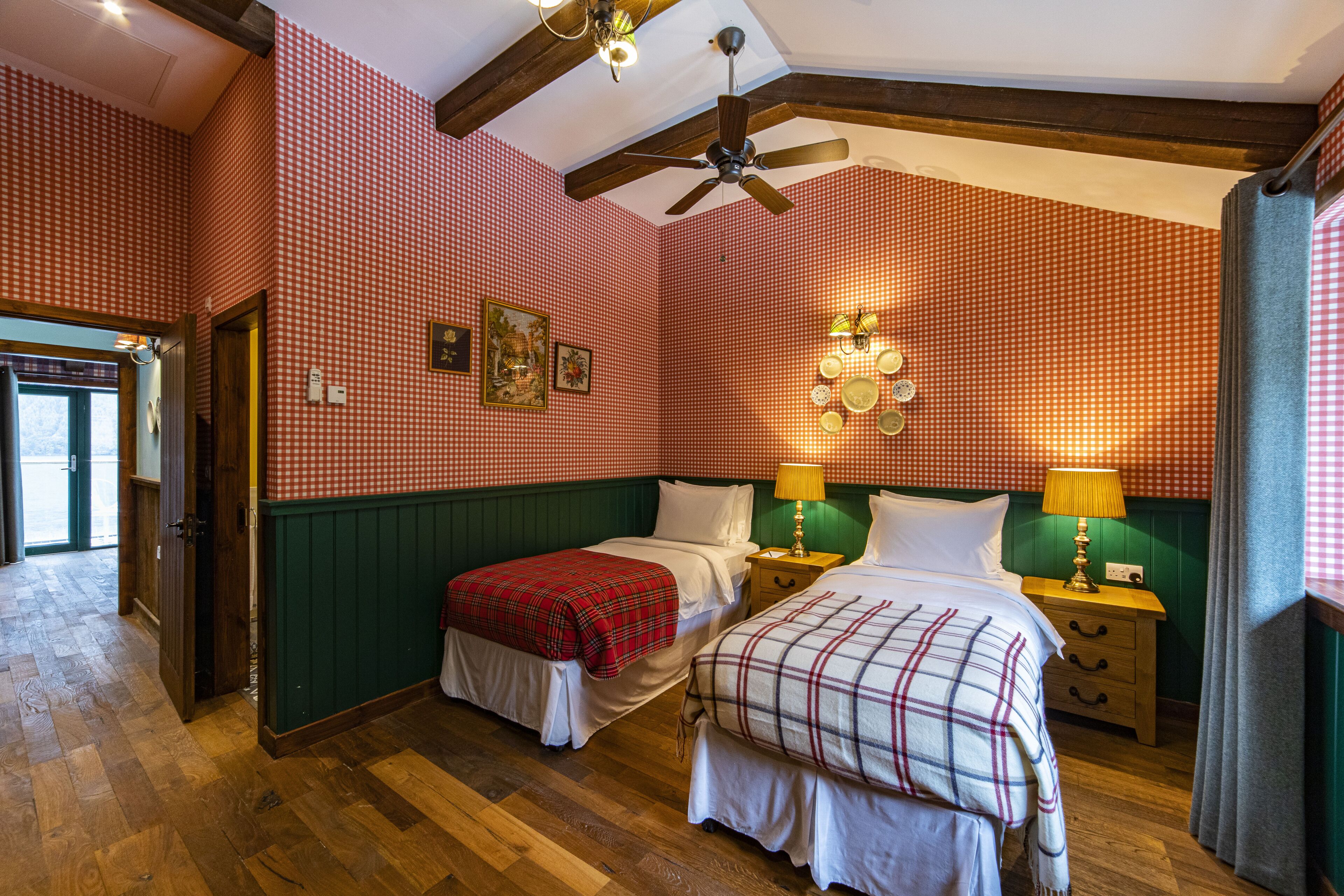Habitación de hotel con dos camas, pisos de madera, ventilador de techo y un estampado de cuadros en la pared.