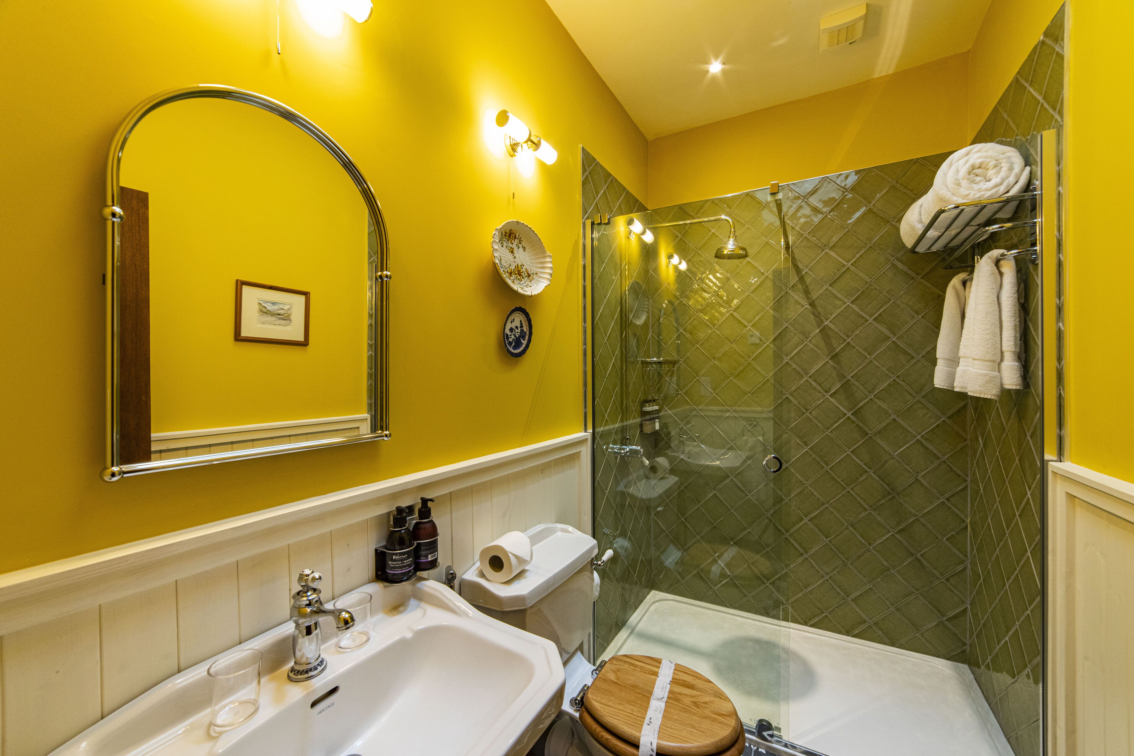 A bathroom with a yellow wall, a glass shower enclosure, a wooden toilet, and a white sink.