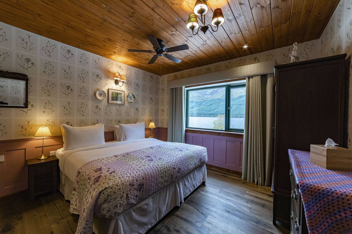 Un dormitorio con una cama grande, un ventilador de techo, una ventana con cortinas y un aparador.