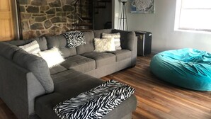 Uma sala de estar acolhedora com um sofá modular cinzento, um puff azul-turquesa, uma parede de pedra, piso de madeira e uma escada.