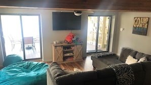 Uma sala de estar com sofá, um bean bag, uma TV fixada na parede e uma porta de vidro deslizante que dá para uma área externa.