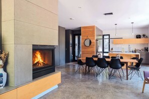 Un salon moderne avec une cheminée, un coin repas avec une table en bois et des chaises noires, et une cuisine avec des meubles en bois et des luminaires suspendus.