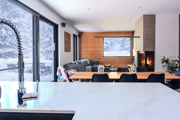 A modern living room with a fireplace, large windows, and a dining area.