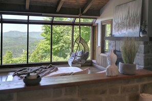 A room with a large window offering a view of a forested area, a hanging chair, and a fireplace with decorative items.