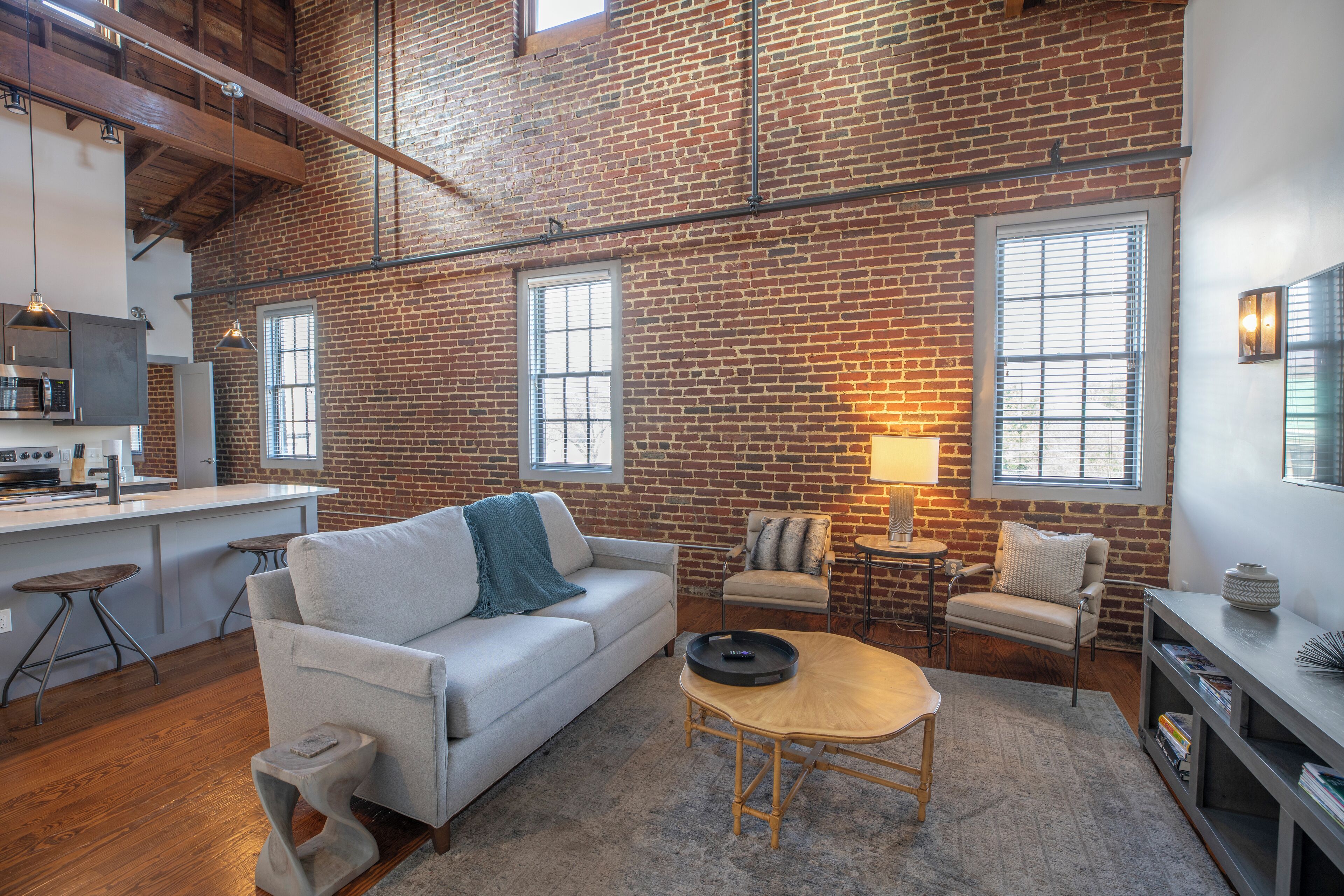 A spacious loft with exposed brick walls, a kitchen area, a seating area with a sofa and chairs, and large windows providing natural light.
