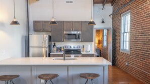 A modern kitchen with stainless steel appliances, a large island with stools, and exposed brick walls. - The Lofts at Downtown Salem (Salem)