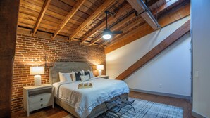 A bedroom with a brick wall, a wooden ceiling, a bed with white bedding, a nightstand, and a floor lamp. - The Lofts at Downtown Salem (Salem)