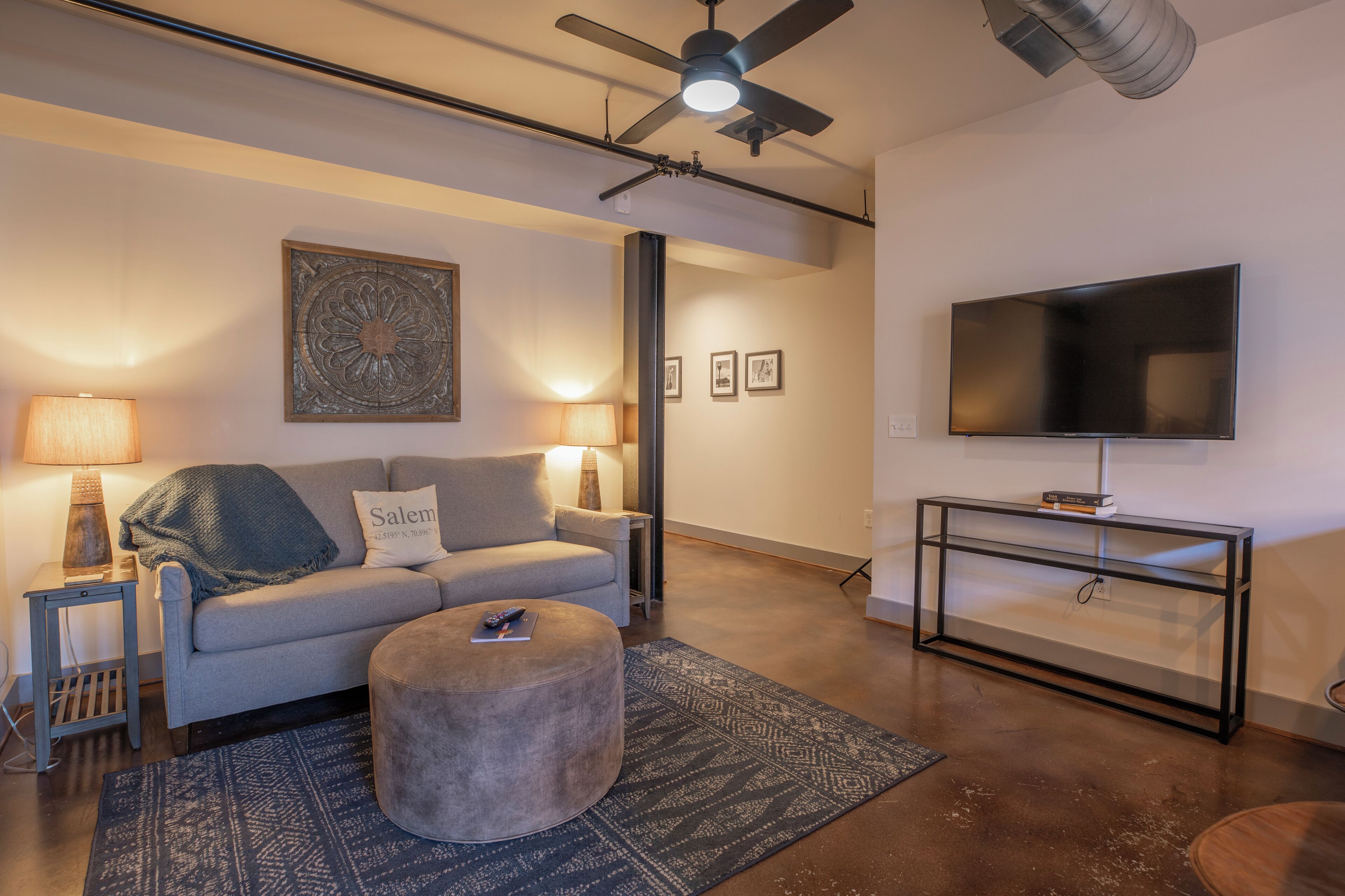 Una sala de estar con un sofá gris, un otoman redondo, un televisor sobre un mueble negro y un ventilador de techo.