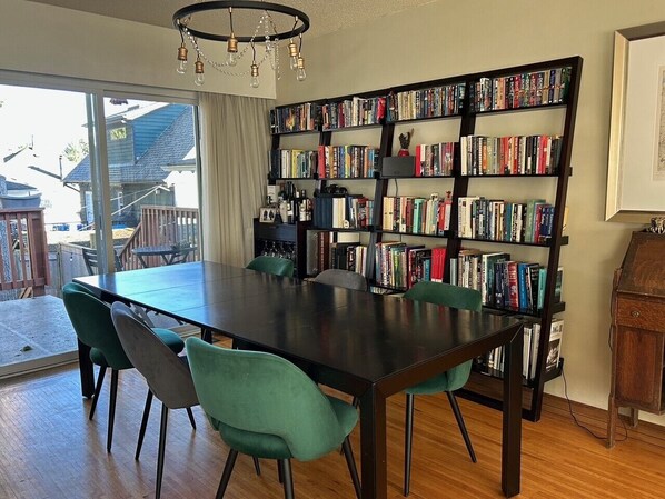 A dining area with a large table and chairs, a bookshelf filled with books, and a view through a glass door to the outside. - Great Family Home in Lynn Valley (North Vancouver)