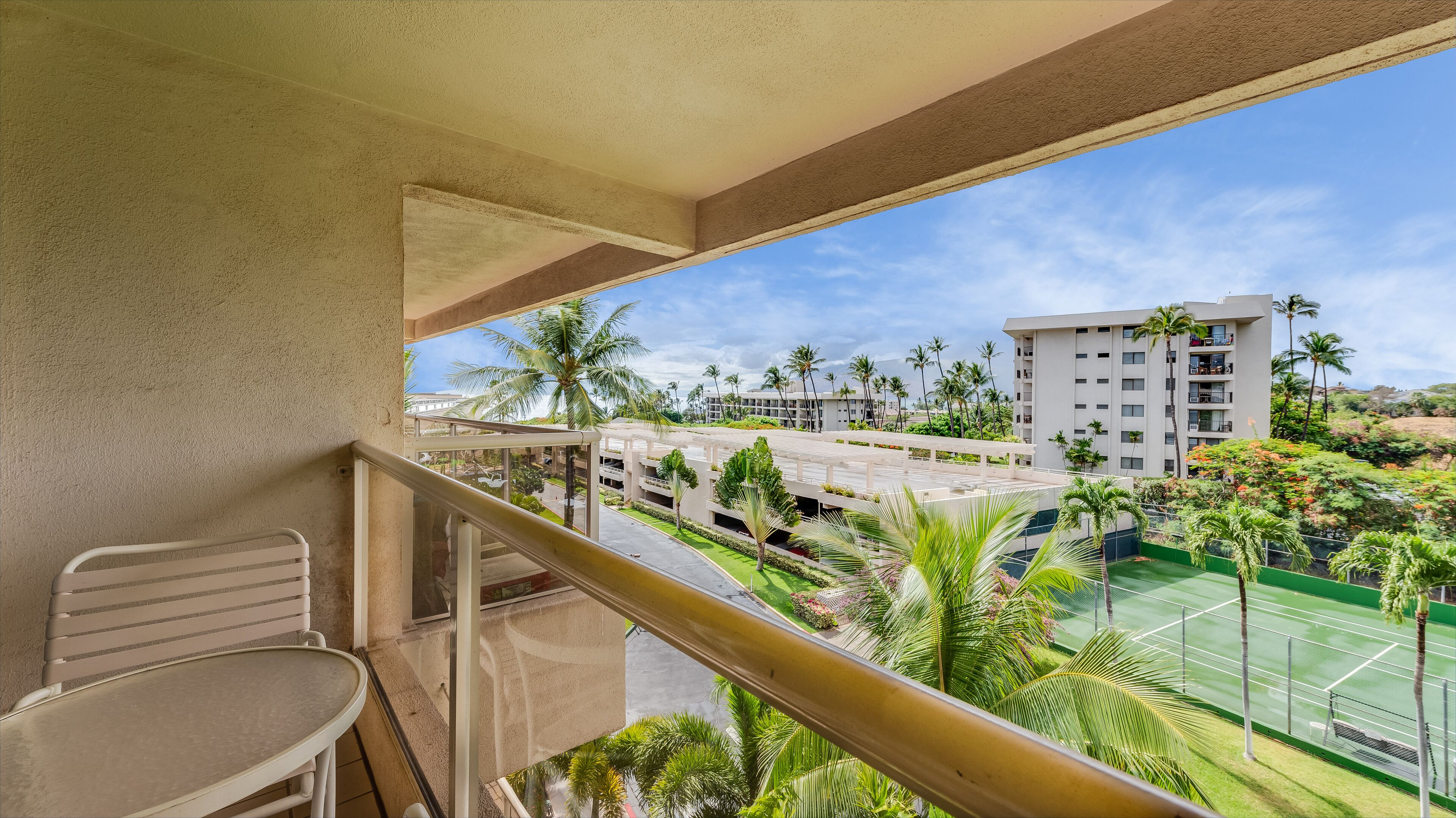 A balcony with a view of a tennis court, palm trees, and a multi-story building.