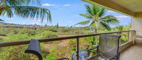 A balcony with two wicker chairs and a glass table, overlooking a lush landscape with palm trees and a distant building complex.