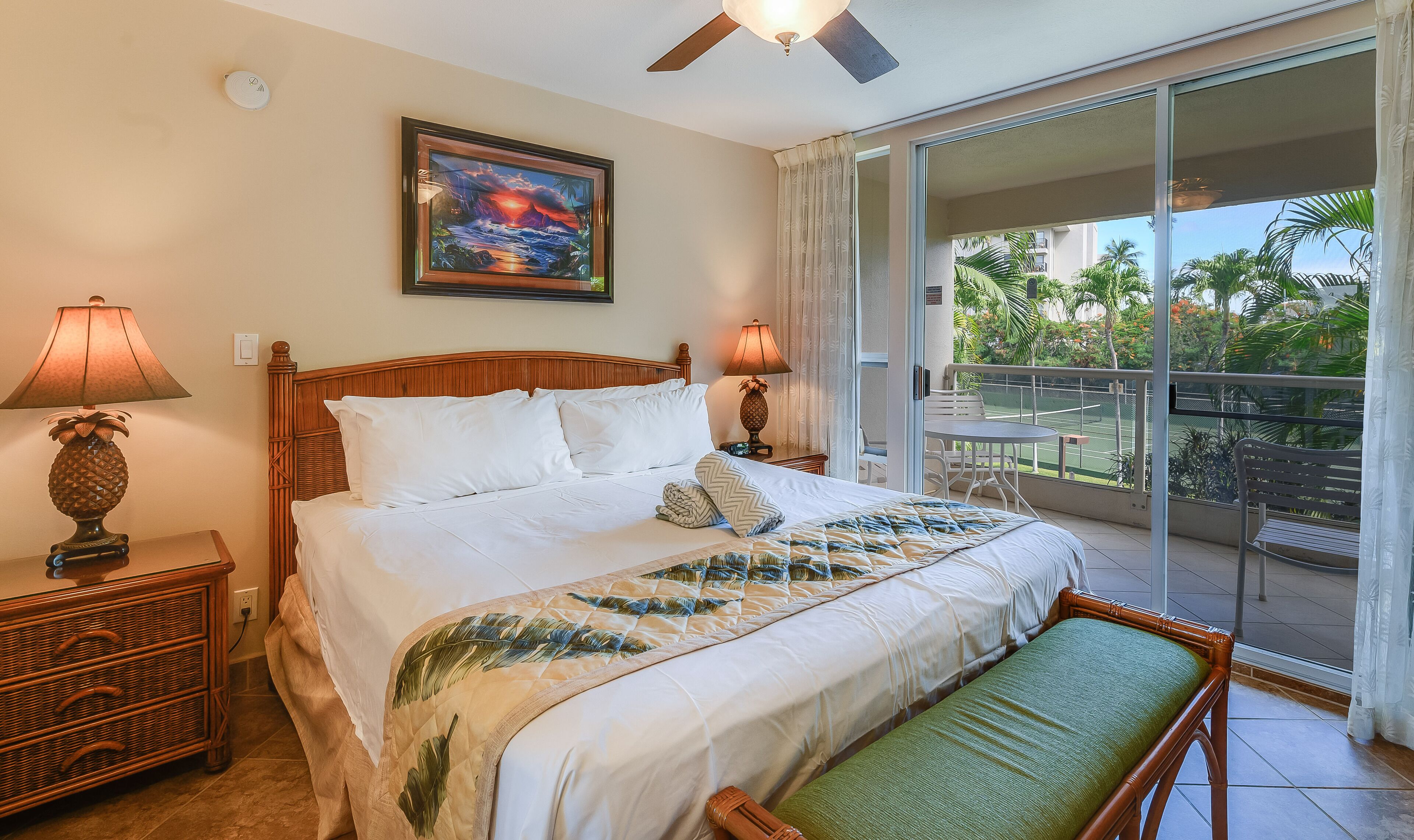 A hotel room with a bed, two bedside lamps, a painting, a balcony with a view of palm trees and a tennis court.