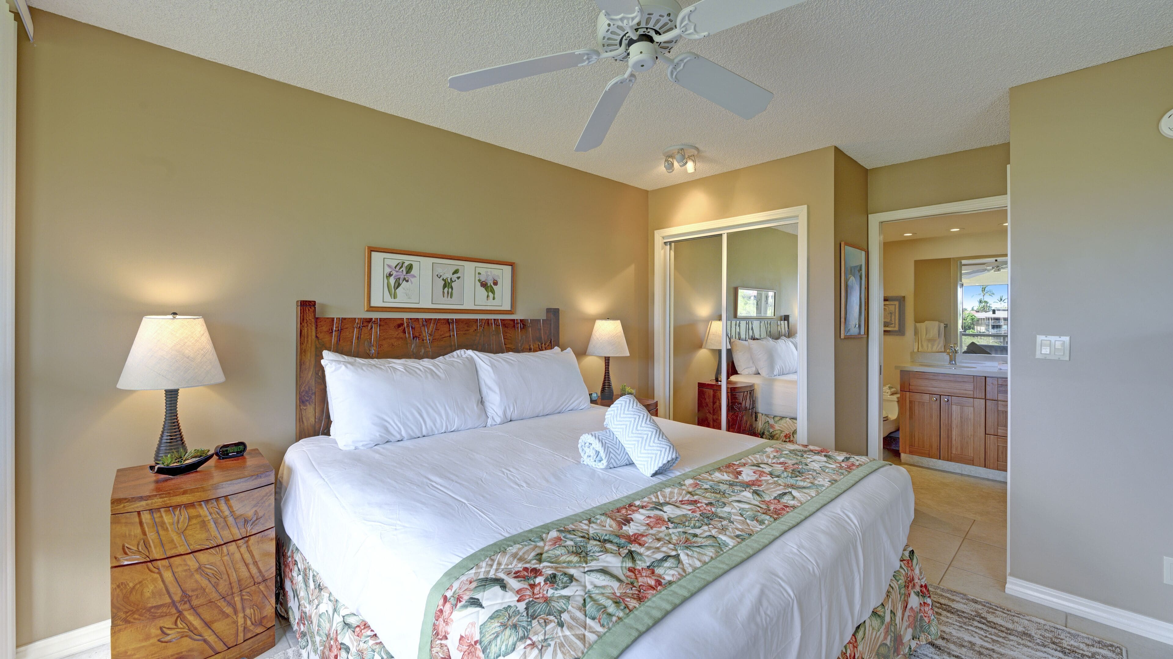 Une chambre d’hôtel avec un lit, deux tables de chevet avec des lampes, un ventilateur de plafond et une salle de bain visible à travers une porte ouverte.