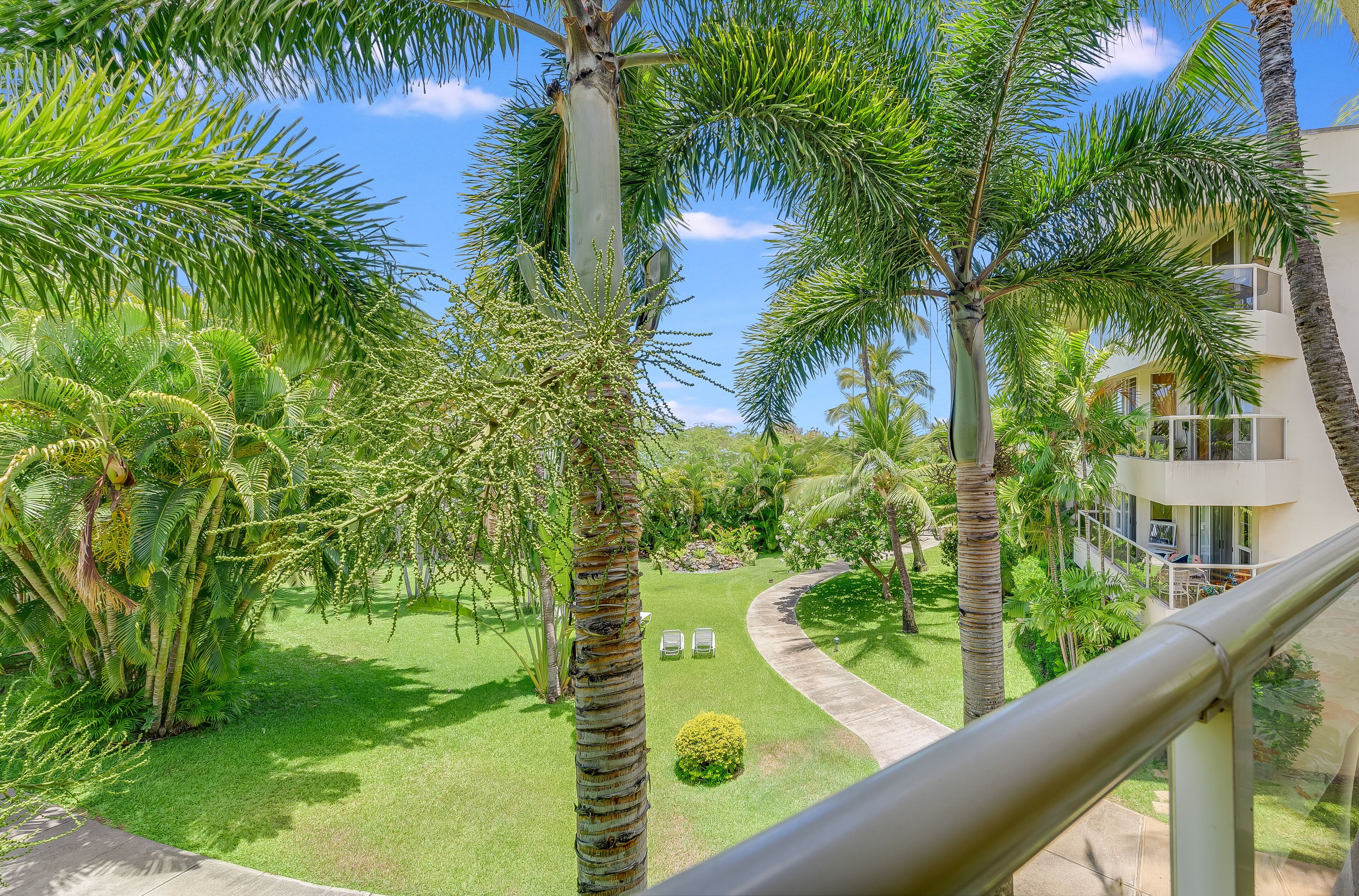 Un jardin luxuriant, agrémenté de palmiers et d’une pelouse bien entretenue, avec un sentier et un bâtiment doté de balcons.