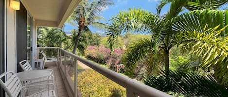 Un balcón con sillas y una mesa blancas, con vistas a un exuberante jardín con diversas plantas y un cielo azul claro.