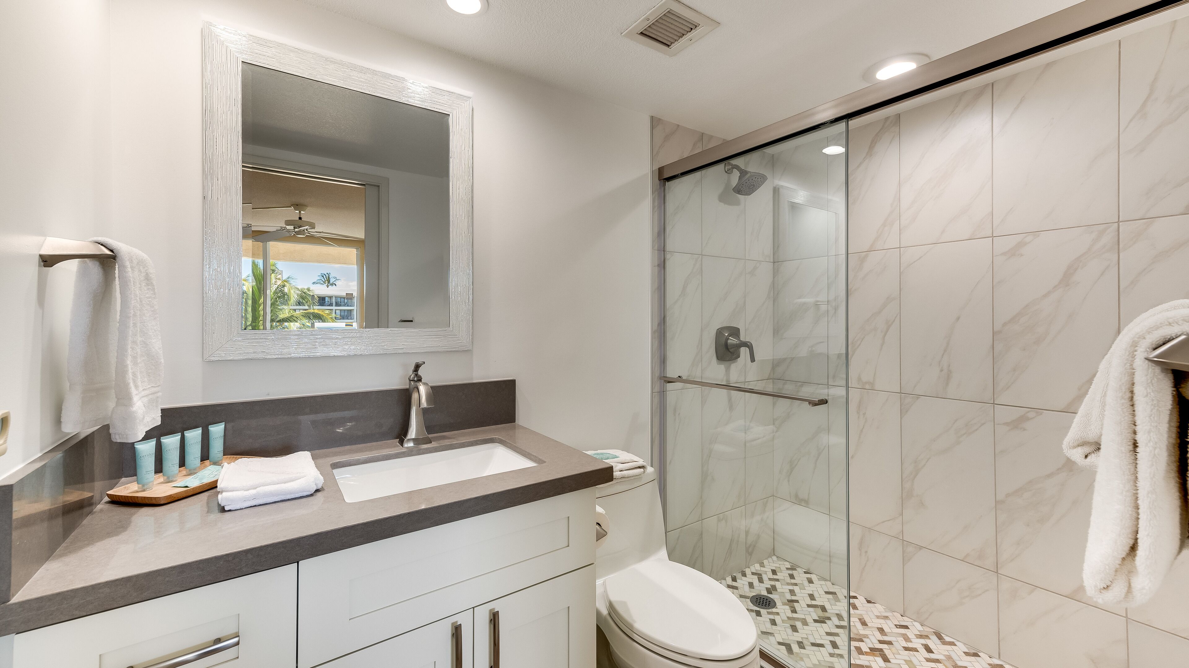 A modern bathroom with a marble shower, a large mirror, and a vanity with toiletries.