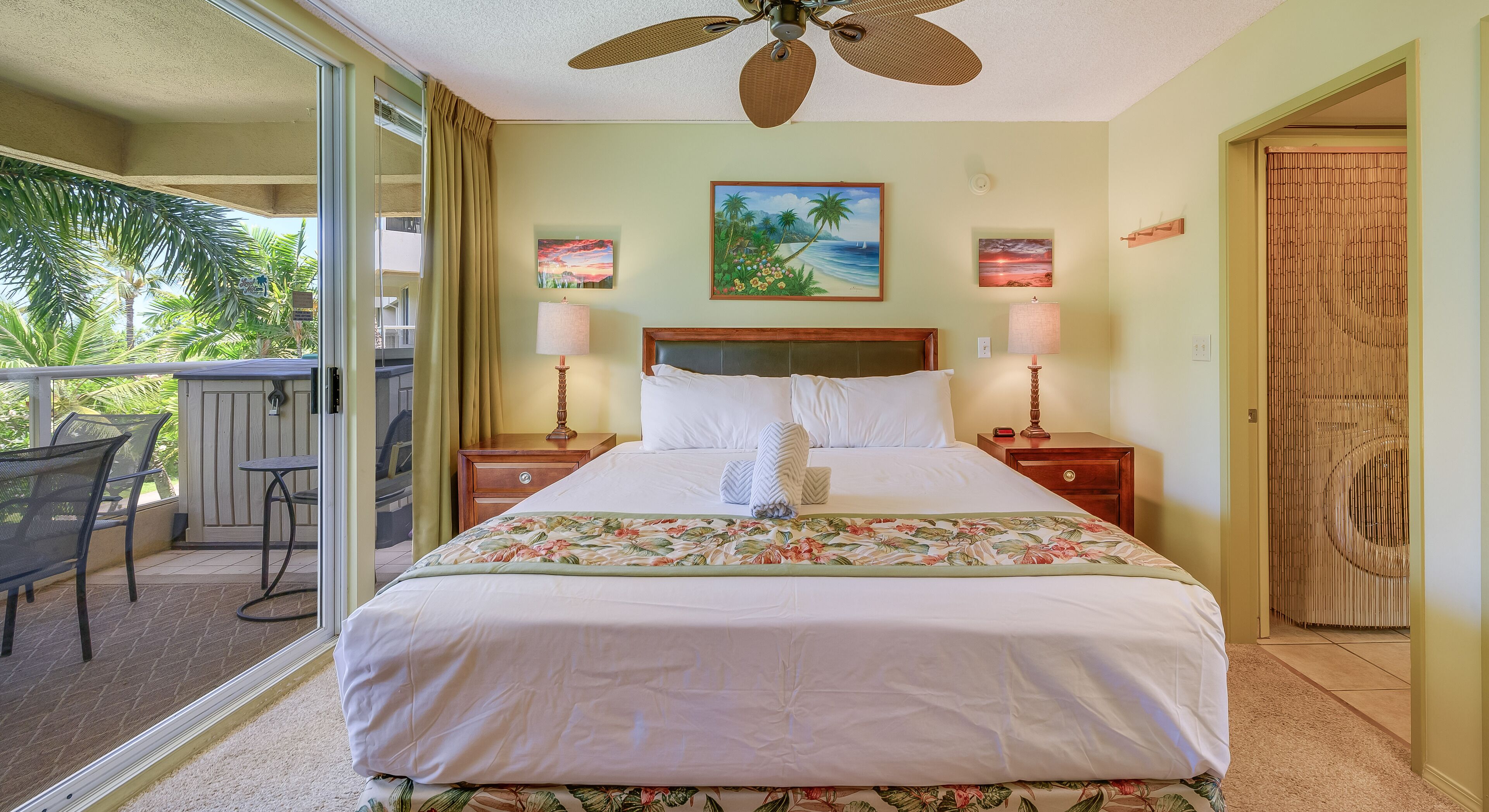 Quarto de hotel com uma cama, duas mesas de cabeceira, um ventilador de teto e uma varanda com mobiliário para exterior.