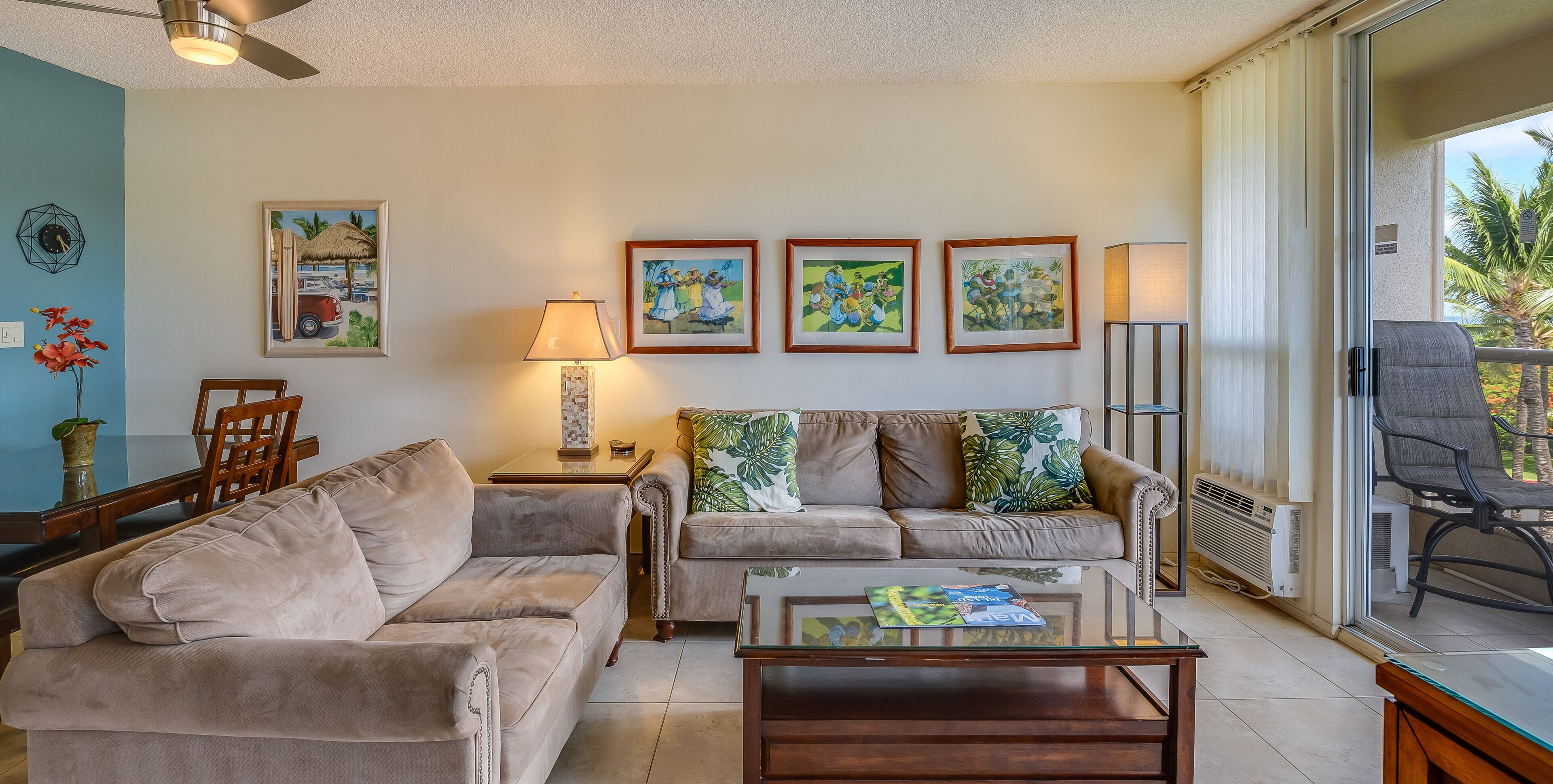 A living room with a beige sofa, a glass coffee table, and a dining table with chairs. There are three framed artworks on the wall, a lamp, and a view of the outdoors through a sliding door.