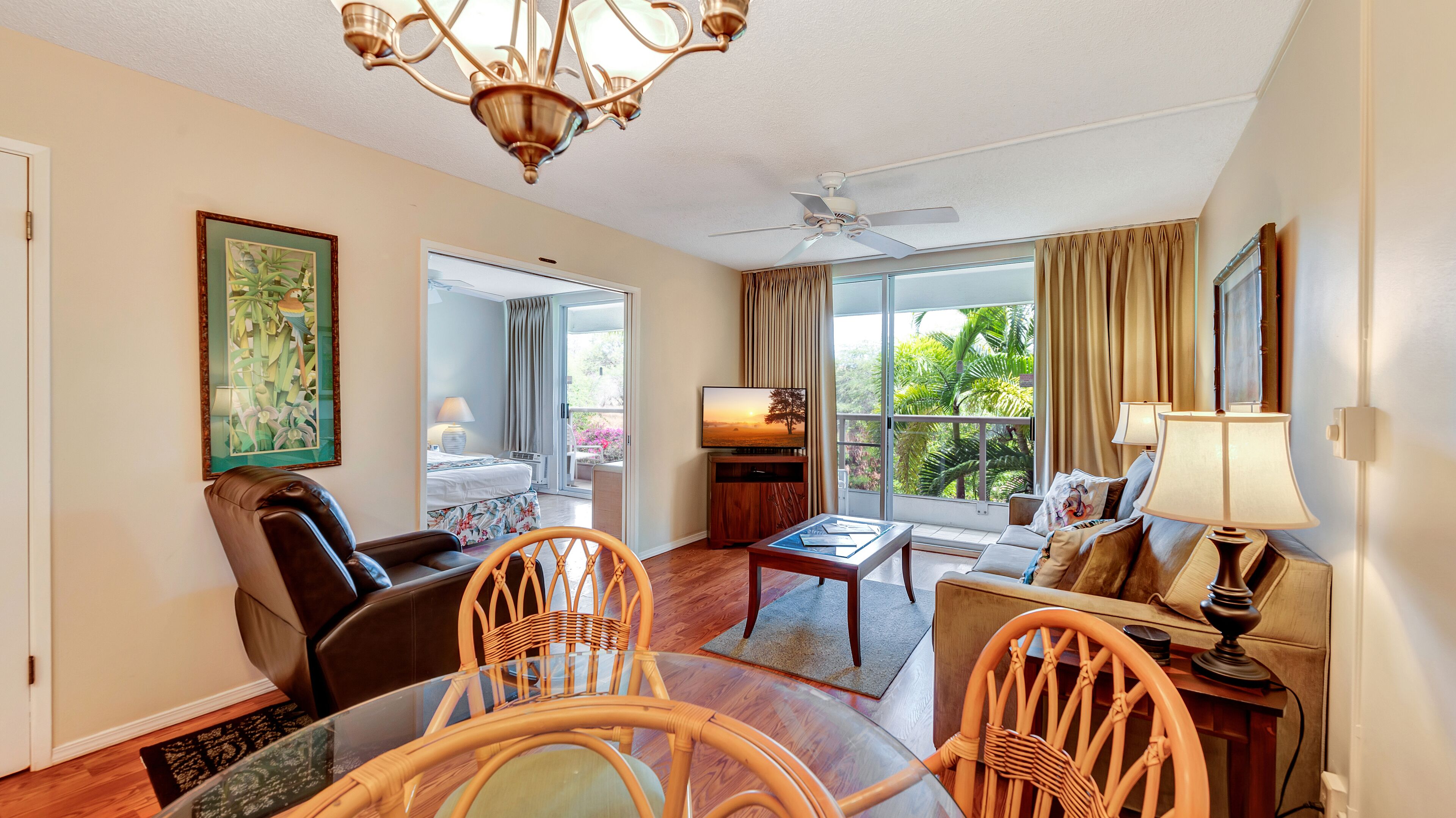 Une chambre d’hôtel avec un balcon, un lit, une télévision, un coin repas avec des chaises et un lustre.