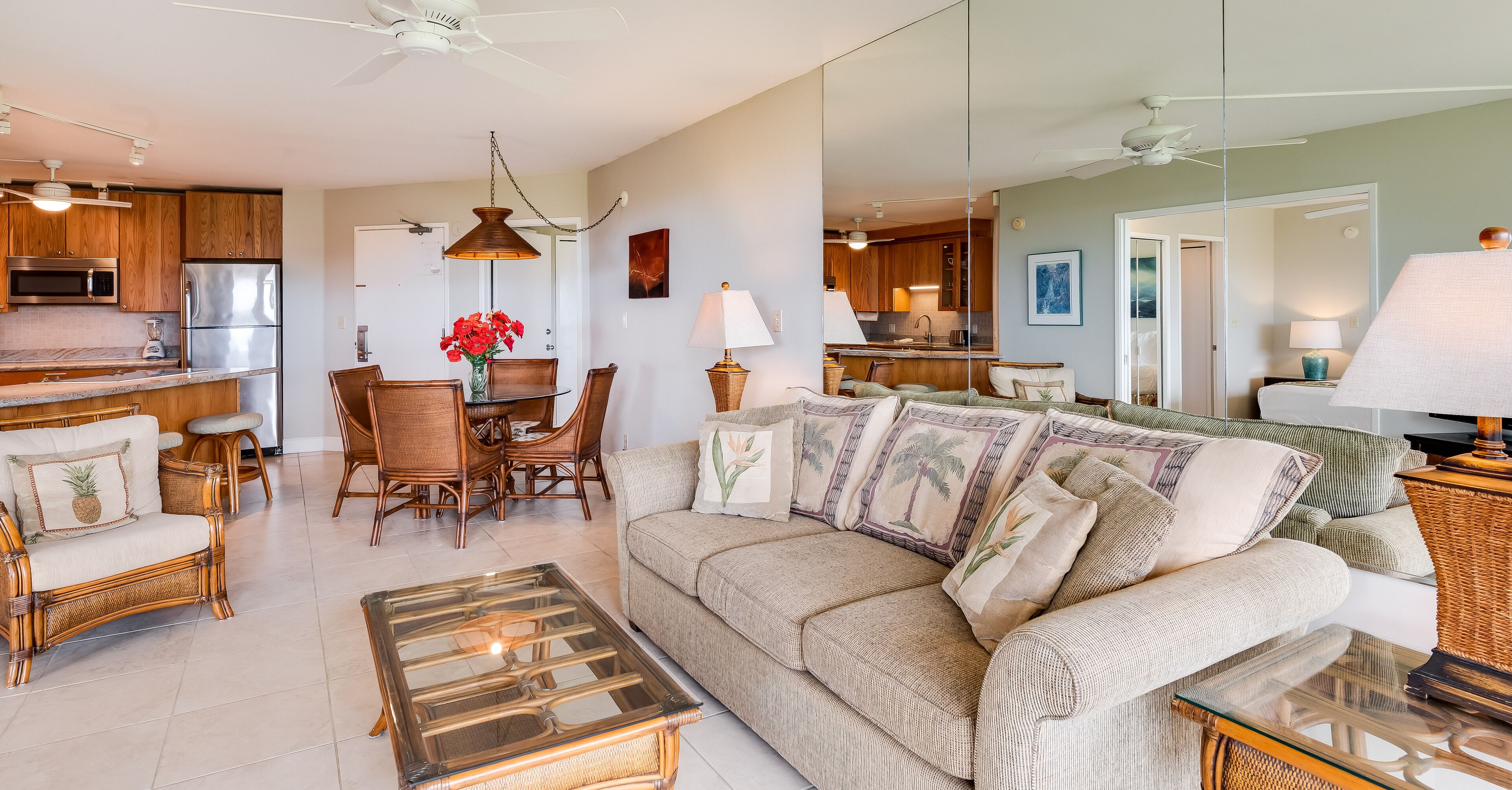 A spacious living room with a large sofa, a glass coffee table, wicker furniture, and a kitchen area with wooden cabinets and modern appliances.