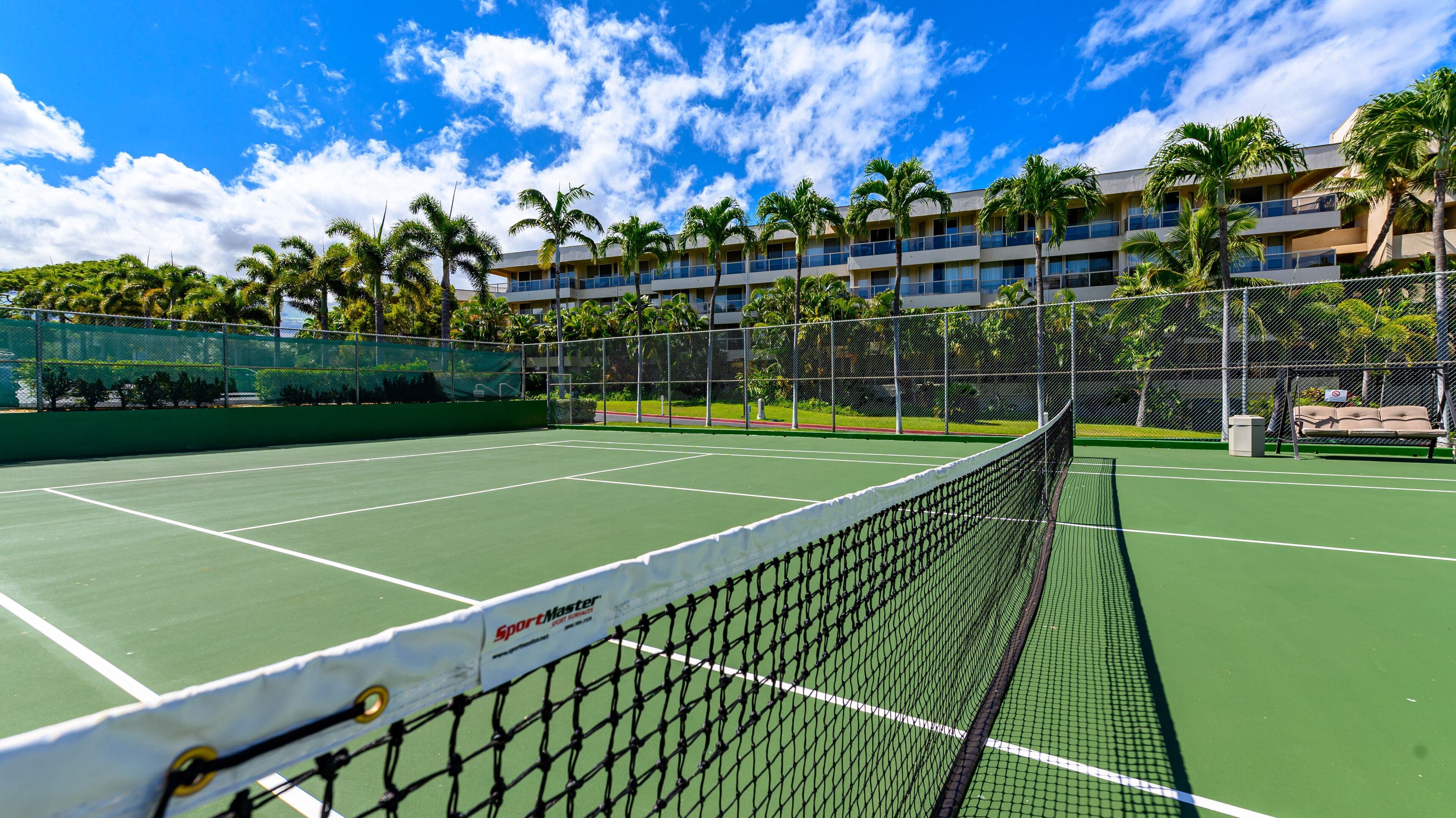 A tennis court with a net, surrounded by a fence and palm trees.