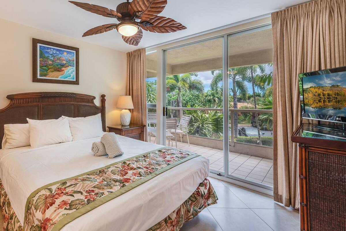 Habitación de hotel con una cama grande, dos lámparas de noche, ventilador de techo, un cuadro, televisor y un balcón con vistas a palmeras y un lago.