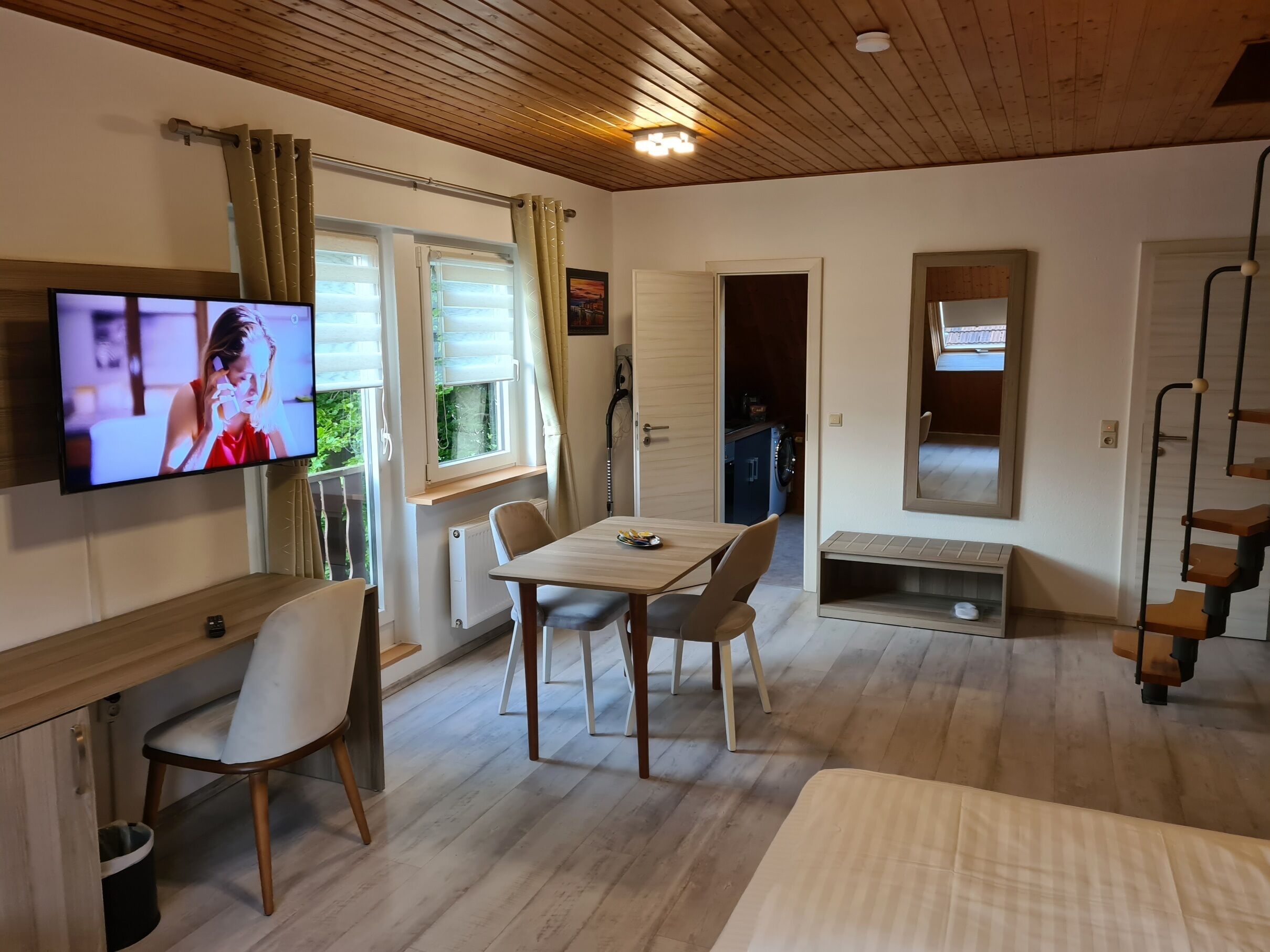 Ein Zimmer mit Holzboden, einem an der Wand befestigten Fernseher, einem Esstisch mit Stühlen, einem Bett und einem Fenster mit Vorhängen.