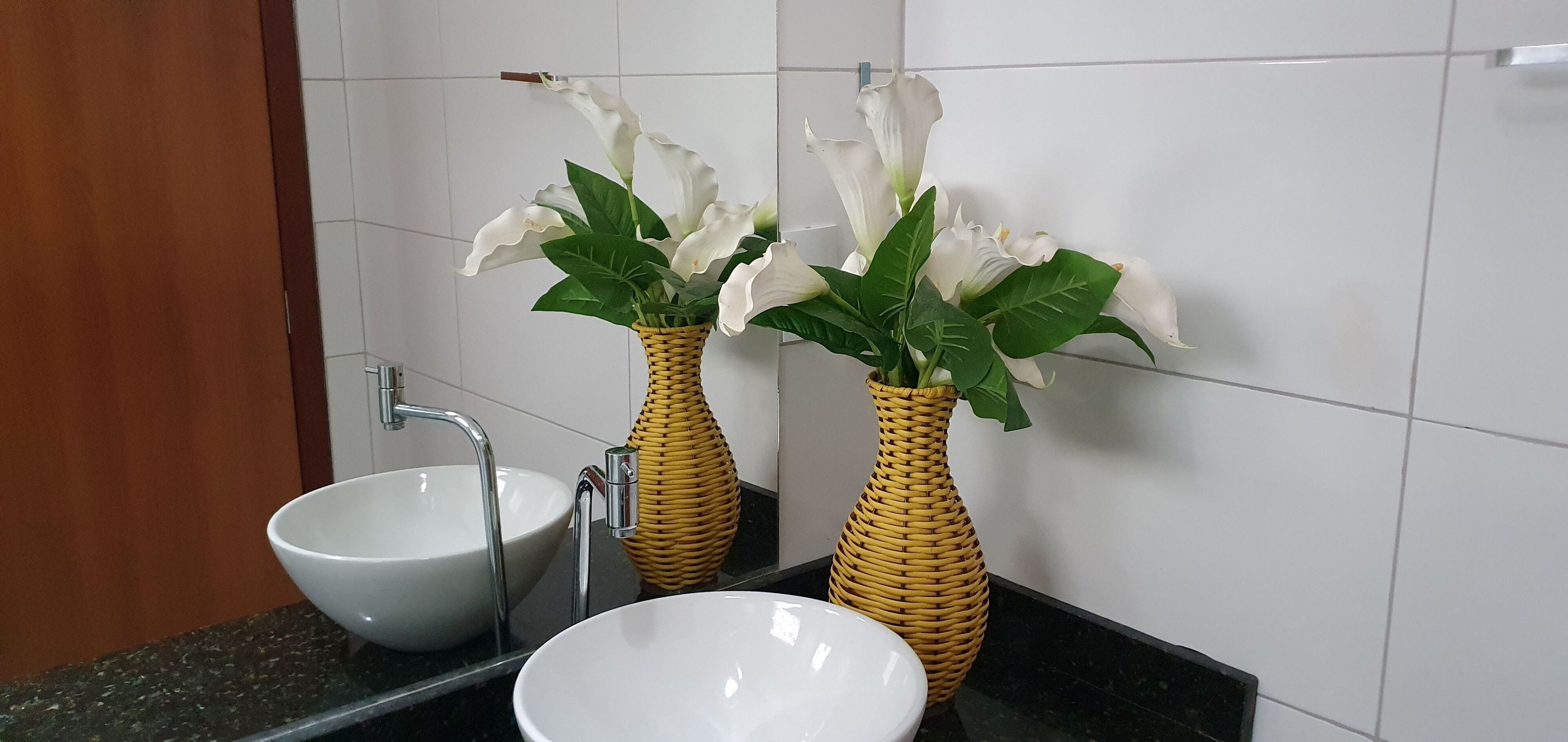 Um banheiro com duas pias de cerâmica branca, um espelho e dois vasos de vime com flores brancas.