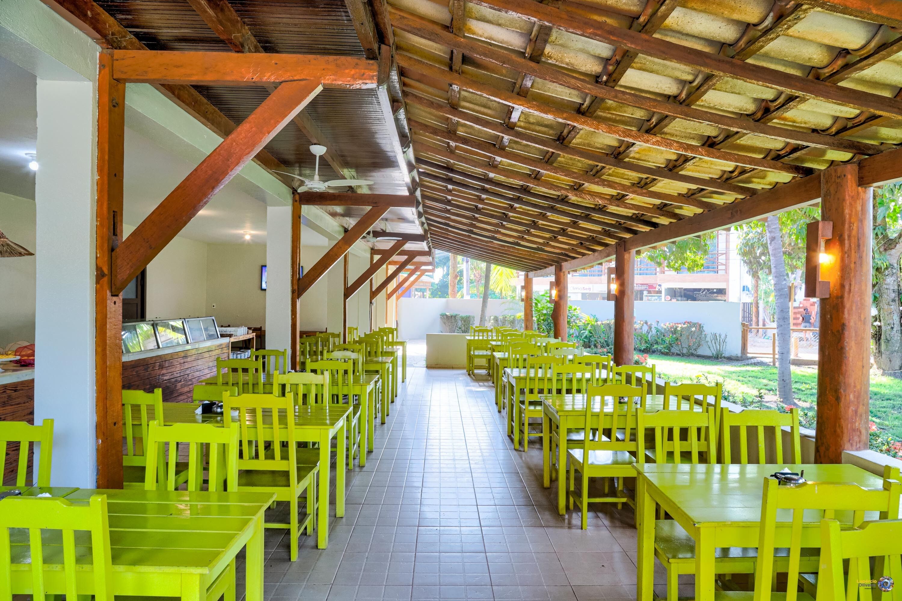 A covered outdoor dining area with yellow tables and chairs, a tiled floor, and a wooden ceiling structure.