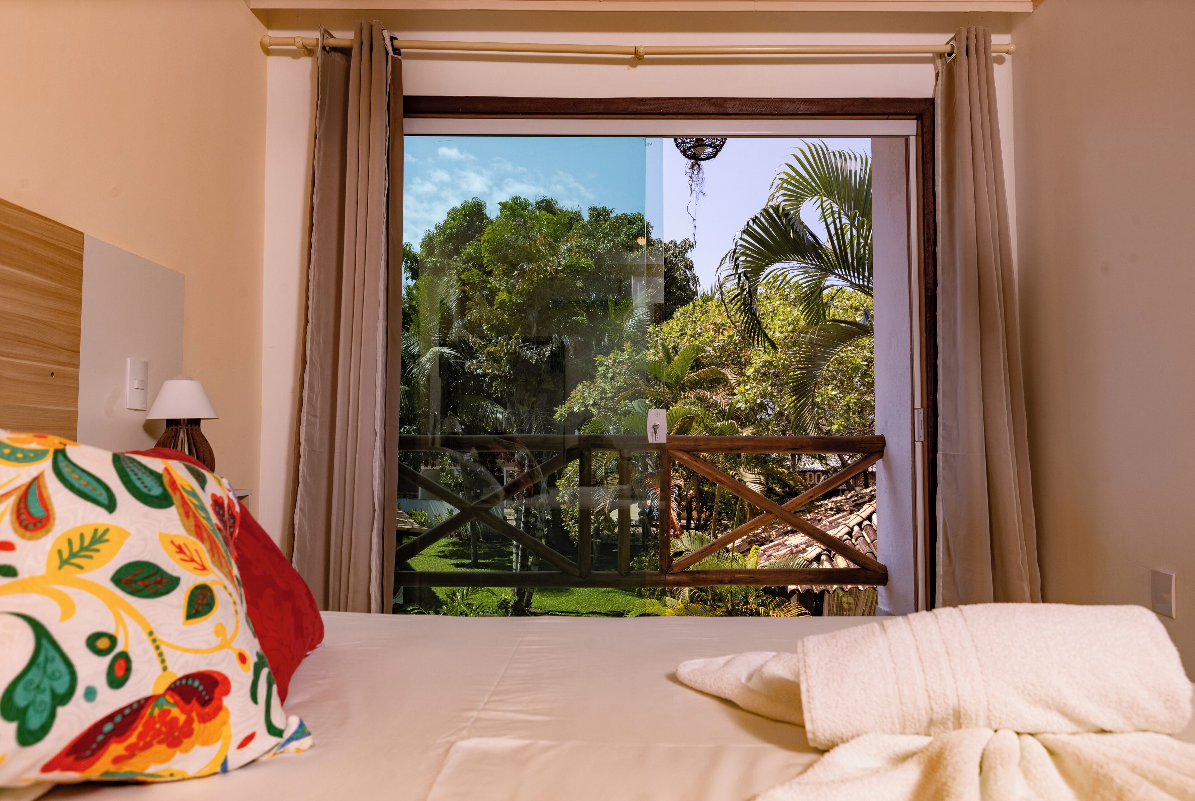 Habitación de hotel con una cama, un cojín colorido, una manta blanca, una mesita de noche con una lámpara y vista a un jardín a través de un ventanal.