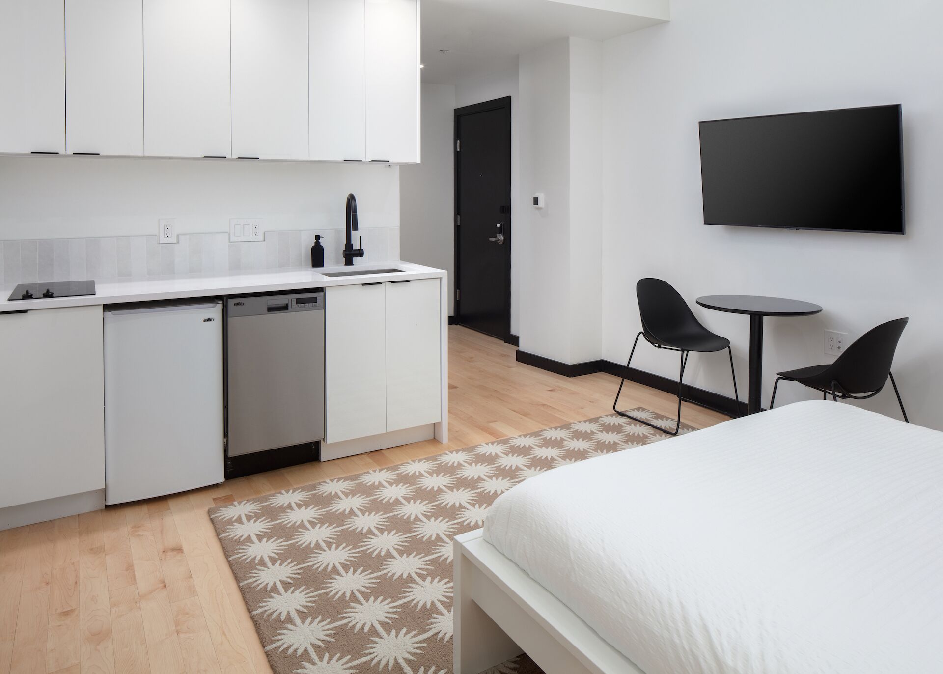 A modern, compact hotel room with a kitchenette, a bed, a small dining area, and a TV.
