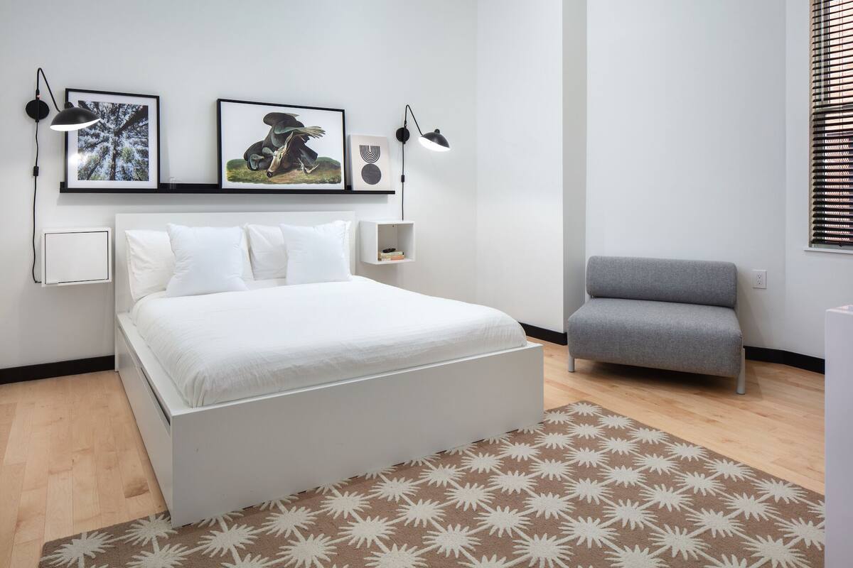 Une chambre moderne avec un grand lit, un fauteuil gris et deux lampes fixées au mur.