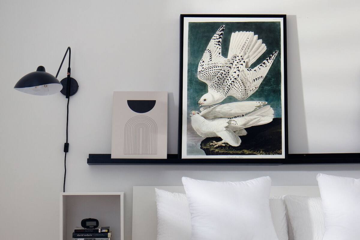 Une chambre avec un lit blanc, une œuvre d’art encadrée représentant un oiseau blanc, une lampe noire et une étagère remplie de livres.