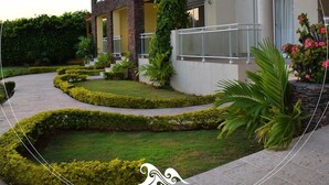 A multi-story building with balconies, a landscaped garden with hedges and plants, and a clear sky. - Wonderful penthouse with pool and sea view (La Romana)