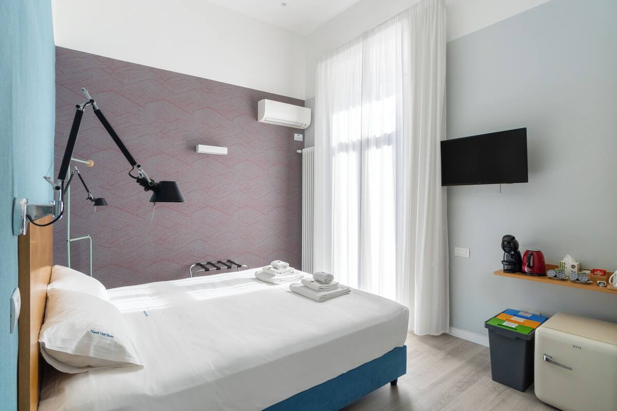 A modern hotel room with a neatly made bed, a desk with a lamp, a TV on the wall, and a small shelf with items.