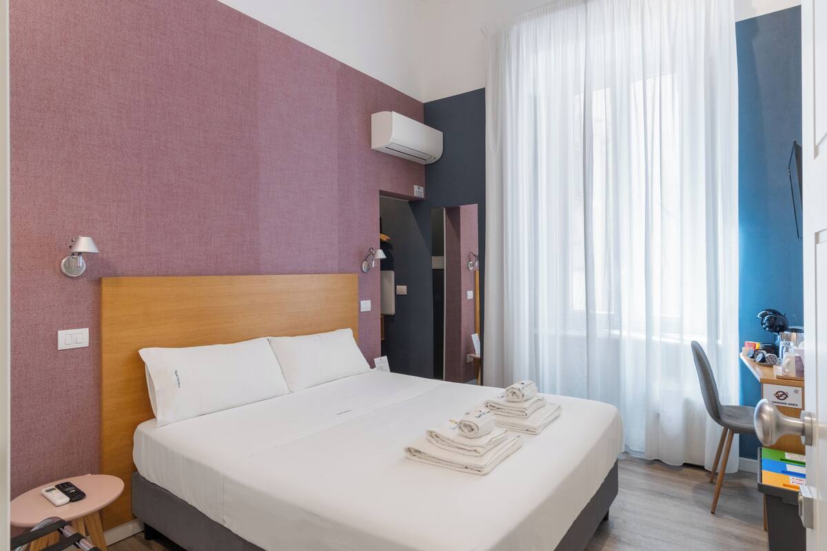 A hotel room with a bed, a desk, a chair, an air conditioner, and a window with curtains.