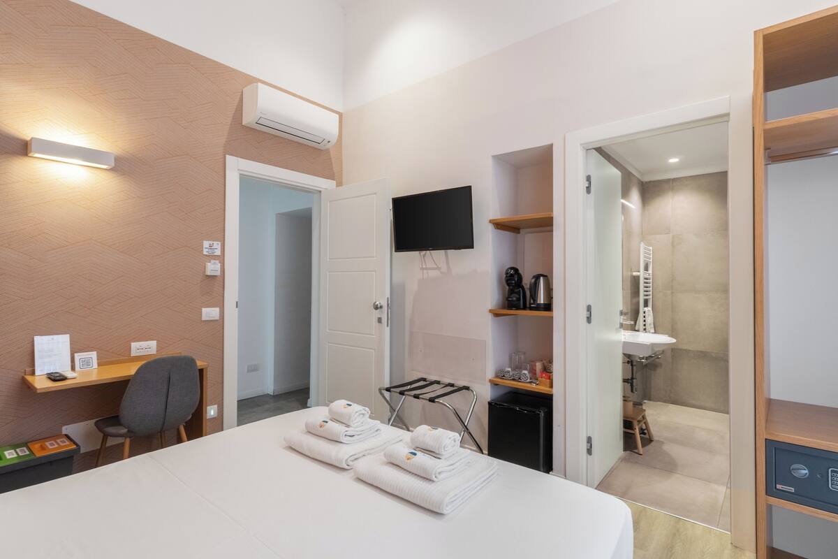 A hotel room with a bed, a desk with a chair, a TV, a shower area, and a small kitchenette.