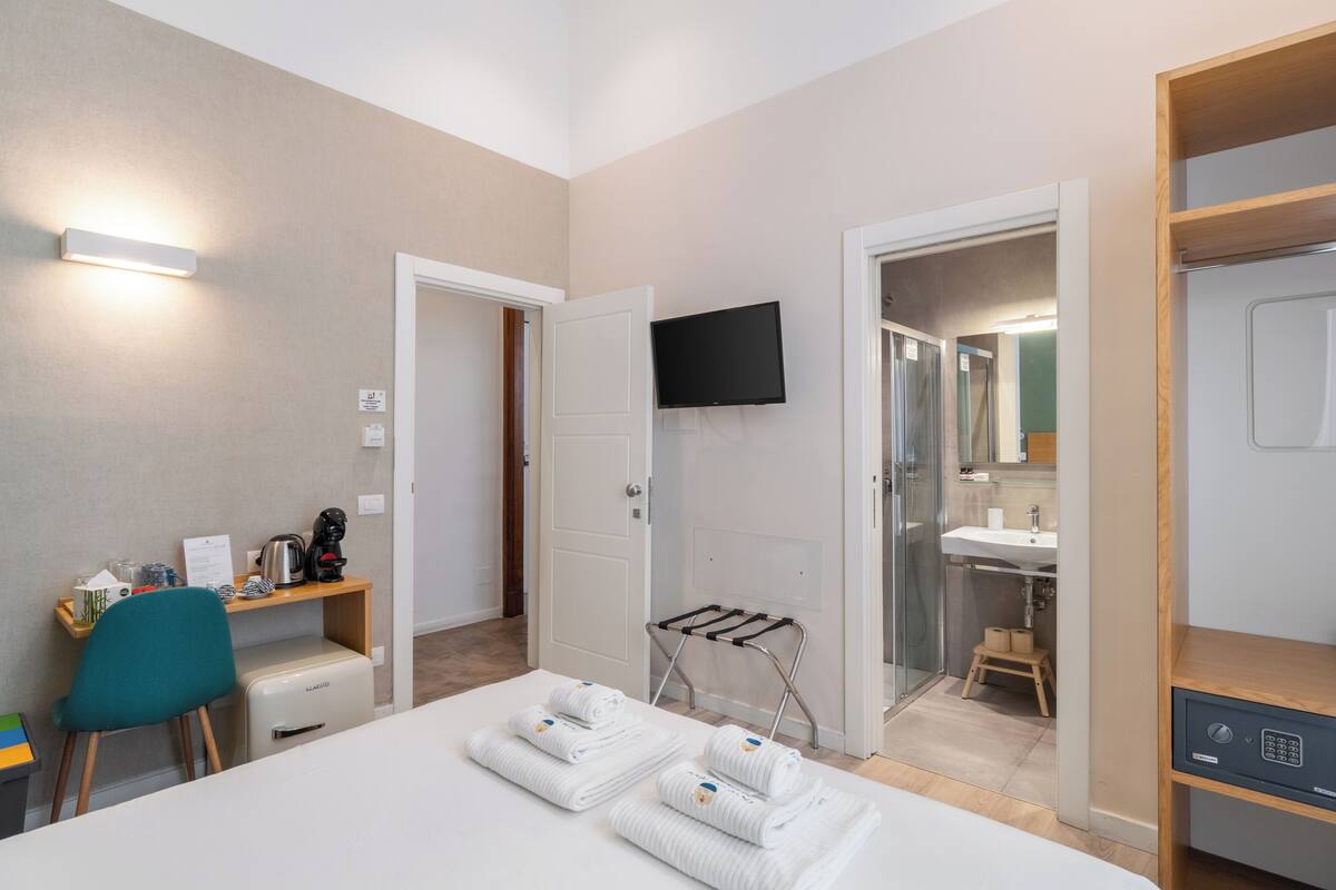 A hotel room with a bed, a desk with a chair, a TV mounted on the wall, a bathroom with a sink and mirror, and a towel rack.