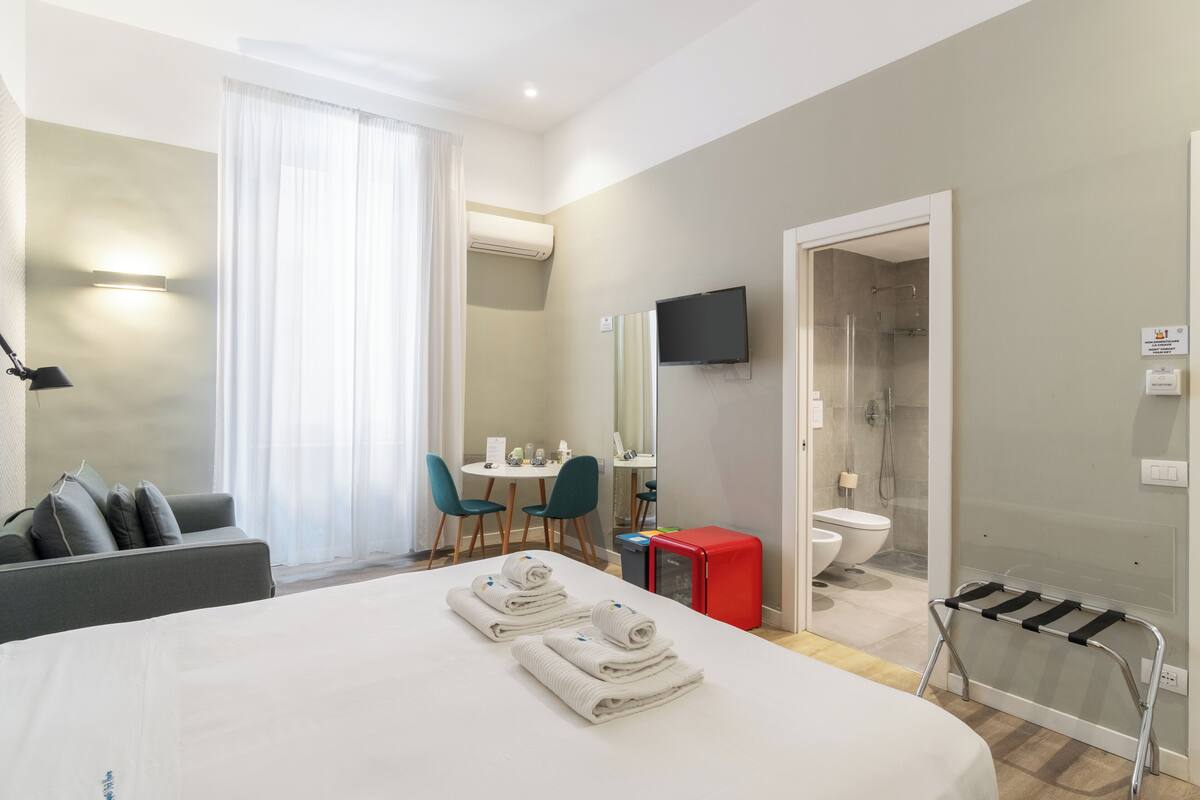 A hotel room with a bed, a desk, a chair, a red cabinet, a TV, a shower, and a towel rack.