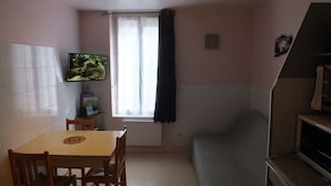 A small, cozy room with a single bed, a table with chairs, a television on the wall, and a window with curtains. - F2 apartment near the spa. (La Bourboule)