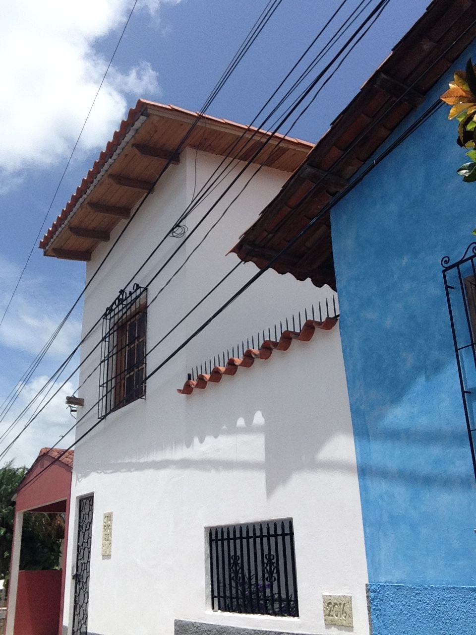 Un edificio de dos pisos con fachada blanca y una sección azul a la derecha. Se observan varias ventanas con barrotes de metal, y una puerta roja es visible a la izquierda.