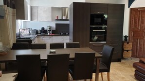 A modern kitchen with dark cabinetry, a dining area with a long table and chairs, and a wooden door. - GITES EN COLLOCATION 33500 NEAC (Néac)