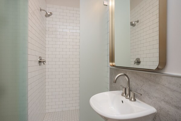 Un baño con paredes de azulejos blancos, un lavamanos blanco con encimera de mármol y una ducha con puerta de vidrio.
