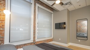 A modern bedroom with a brick wall, a large window with blinds, a flat-screen TV, and a mirror on the wall. - The Record Room -steps to Broadway & Printer’s Alley (Nashville)