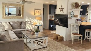 Un salon comprenant un canapé, une table basse et un téléviseur. Il y a également un coin cuisine avec un réfrigérateur, un four à micro-ondes et un îlot central en bois.