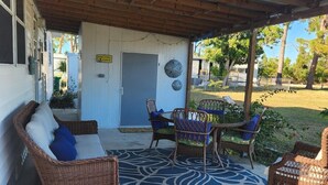 Une terrasse couverte, meublée de meubles en osier, agrémentée d’un tapis à motifs et dotée d’une porte sur laquelle est apposée une pancarte.
