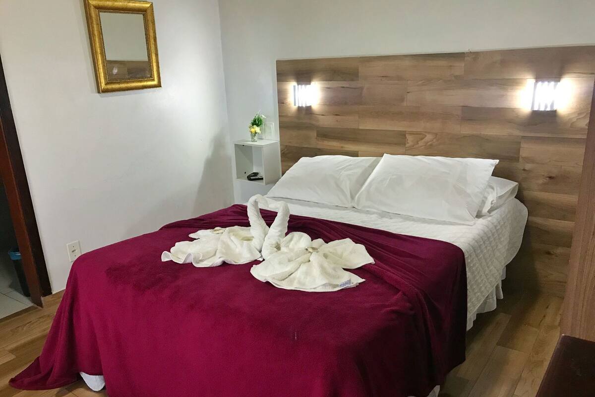 A hotel room with a wooden headboard, a bed with white linens, a red bedspread, and two white towels arranged on the bed.